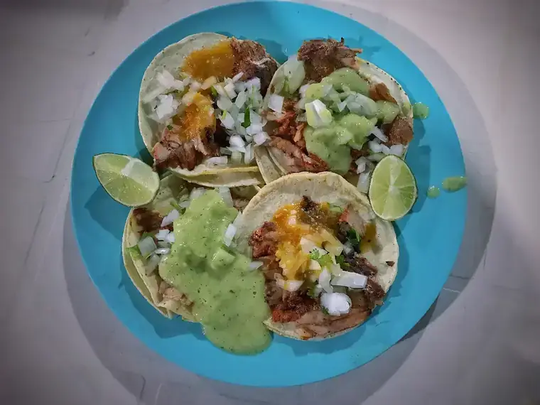 Cuatro tacos al pastor en un plato azul con salsa verde y limón