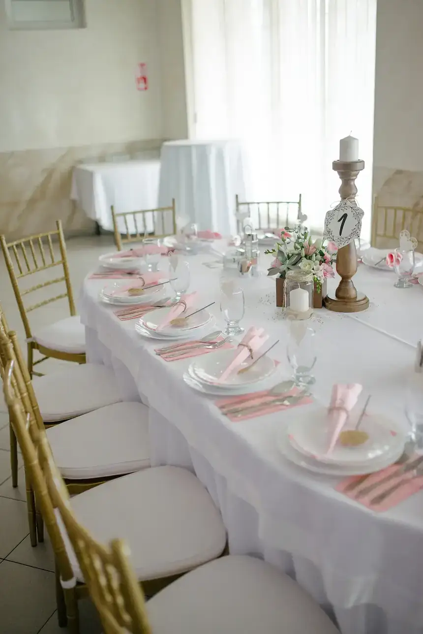 Mesa larga vestida de blanco con sillas doradas, servilletas rosas y centro de flores